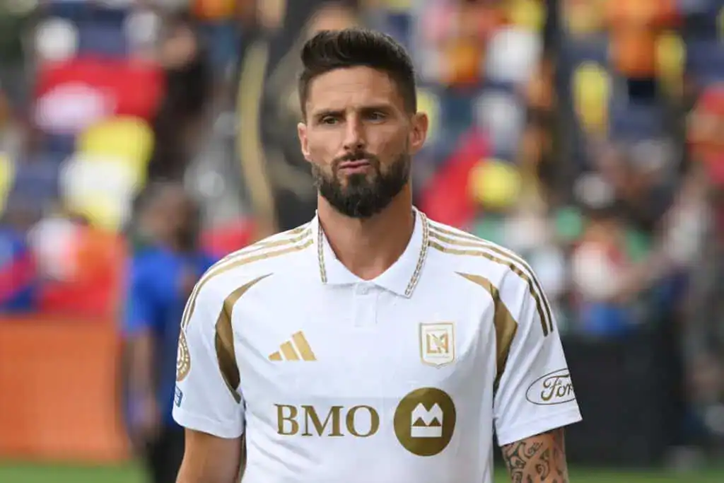 A soccer player in a white jersey with gold accents stands on the field, surrounded by a colorful crowd in the background.