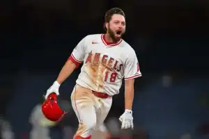 A player in a stained Los Angeles Angels uniform walks confidently on the field, holding a red helmet, amidst a blurred stadium backdrop.