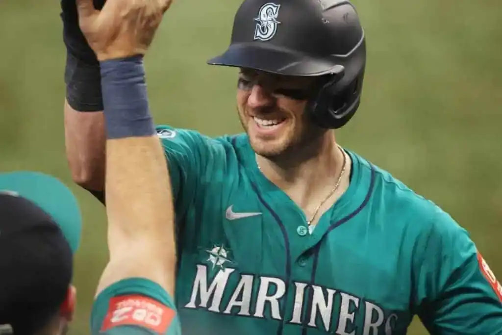 A baseball player in a teal Mariners jersey celebrates with a teammate, raising his arm in joy during a game.