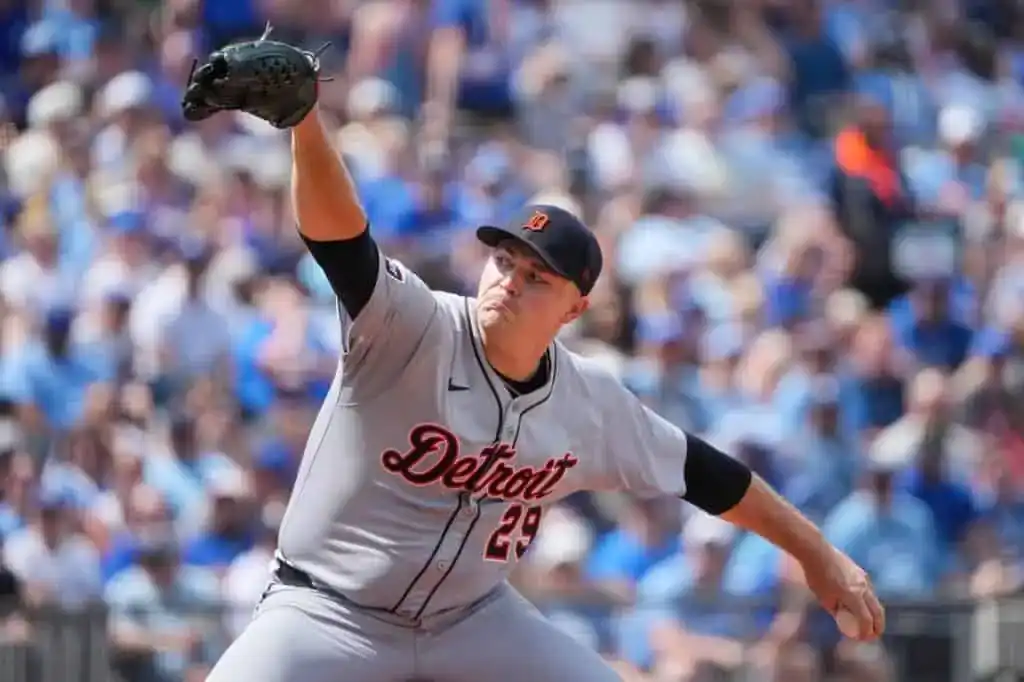 A baseball pitcher in a gray Detroit Tigers uniform winds up to throw against a blurred crowd in the background during a sunny game.