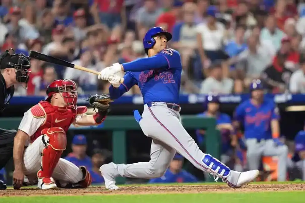 A baseball player swings at a pitch in a packed stadium, with the catcher poised in red gear behind him.