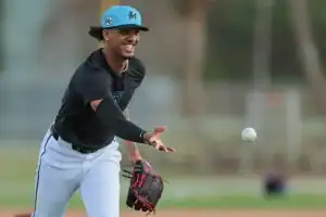 A player wearing a black shirt and white pants prepares to catch a baseball during practice, with palm trees in the background.