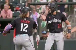 Two baseball players in black jerseys celebrate in a stadium, one with "BADER" on the back, while the crowd cheers in the background.