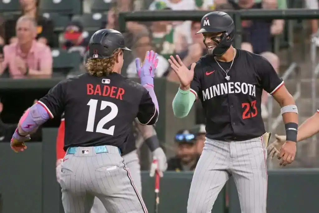Two baseball players in black jerseys celebrate in a stadium, one with "BADER" on the back, while the crowd cheers in the background.