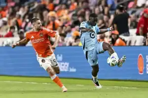 A soccer player in a sky-blue uniform skillfully controls the ball while an opponent in an orange uniform trails behind.