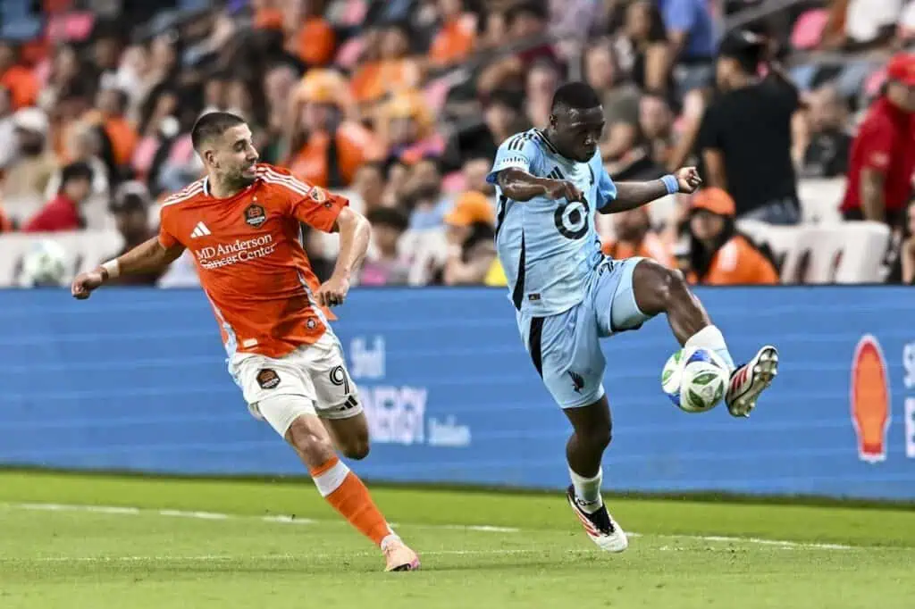 A soccer player in a sky-blue uniform skillfully controls the ball while an opponent in an orange uniform trails behind.
