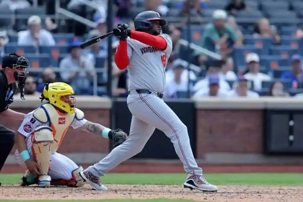 A baseball player in a gray Washington jersey swings at a pitch while a catcher in a yellow helmet crouches behind home plate.