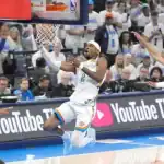 A basketball player in a white and blue jersey attempts a layup while being guarded by an opponent during a dynamic game scene.