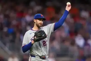 New York Mets player Pete Alonso stands on the pitcher's mound wearing a grey road jersey and blue cap. He is holding up a baseball in his right hand and wearing a glove on his left.