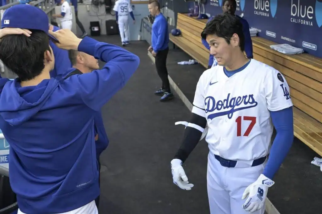 Two baseball players in Dodgers uniforms chat in the dugout, with one wearing a white jersey and the other in a blue hoodie.