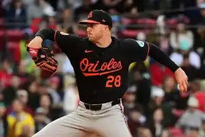 A baseball pitcher in an orange and black Orioles jersey winds up for a pitch on a field filled with spectators in the background.