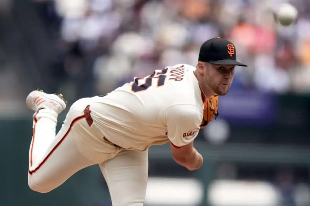San Francisco Giants baseball pitcher in a cream uniform and black cap mid-throw during a game, with his arm extended and leg raised, focused on delivering the pitch.