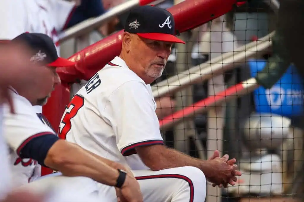 Atlanta Braves baseball coach in a white uniform and black-and-red cap sitting in the dugout during a game, looking toward the field.