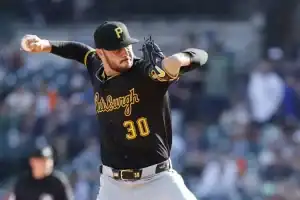 A pitcher in a black Pittsburgh Pirates jersey winds up to throw a baseball, with a blurred crowd in the background.