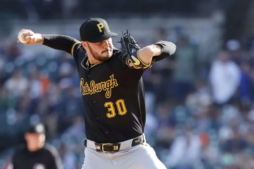 A pitcher in a black Pittsburgh Pirates jersey winds up to throw a baseball, with a blurred crowd in the background.