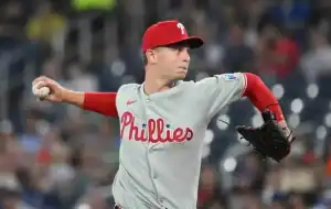 A Philadelphia Phillies pitcher in a gray jersey and red cap throws a baseball during a game, with fans blurred in the background.