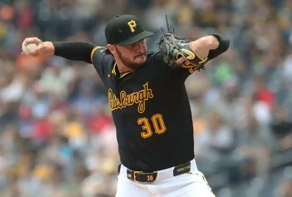 A pitcher in a black Pittsburgh Pirates jersey prepares to throw a baseball during a game, with fans blurred in the background.