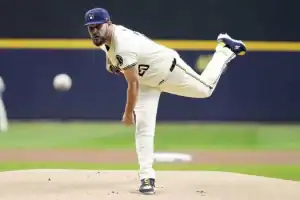 A pitcher in a cream baseball uniform winds up on the mound, preparing to throw a pitch, with the ball in mid-air.
