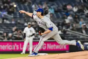 A Texas Rangers pitcher delivers a pitch on the mound, with players and a crowd visible in the stadium background.