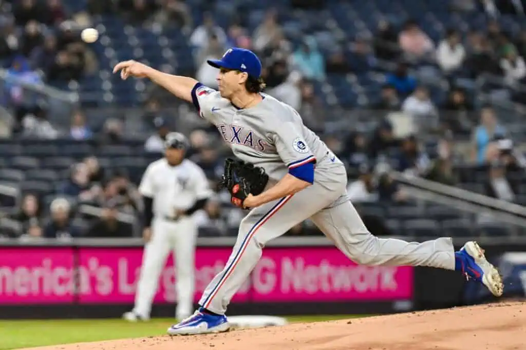 A Texas Rangers pitcher delivers a pitch on the mound, with players and a crowd visible in the stadium background.