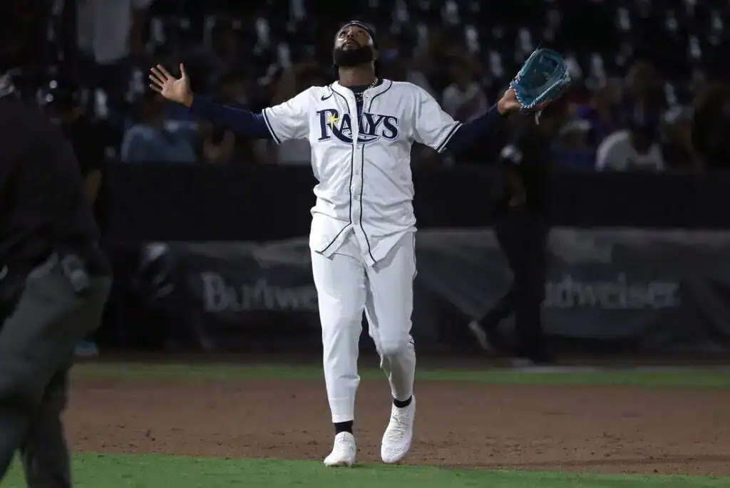 A baseball player in a Rays uniform stands on the field, arms outstretched in celebration or excitement, under stadium lights.