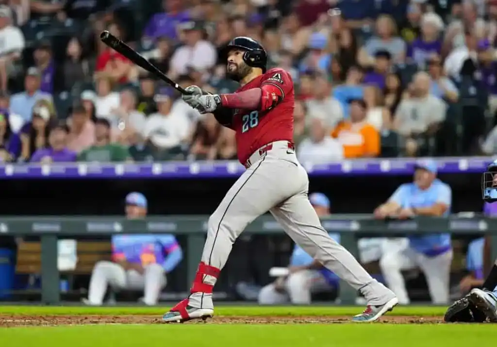 A baseball player in a red jersey swings a bat during a game, with spectators visible in the blurred background.