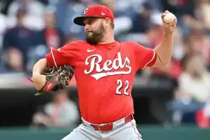 A Cincinnati Reds pitcher in a red jersey (#22) prepares to throw a baseball during a game, with a blurred crowd in the background.