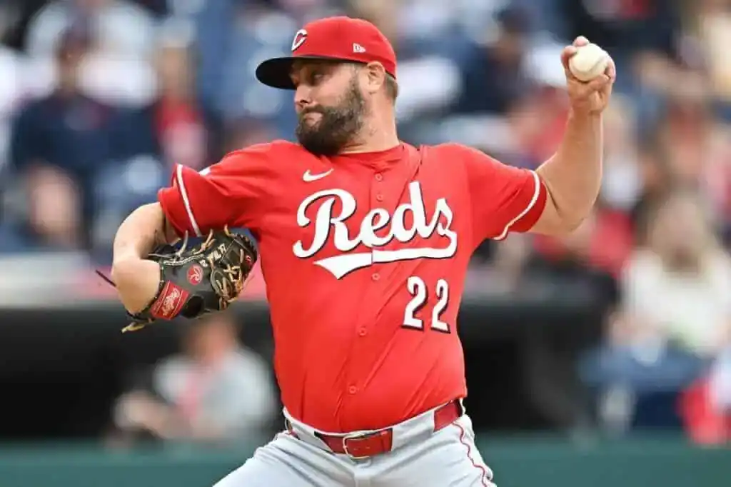 A Cincinnati Reds pitcher in a red jersey (#22) prepares to throw a baseball during a game, with a blurred crowd in the background.