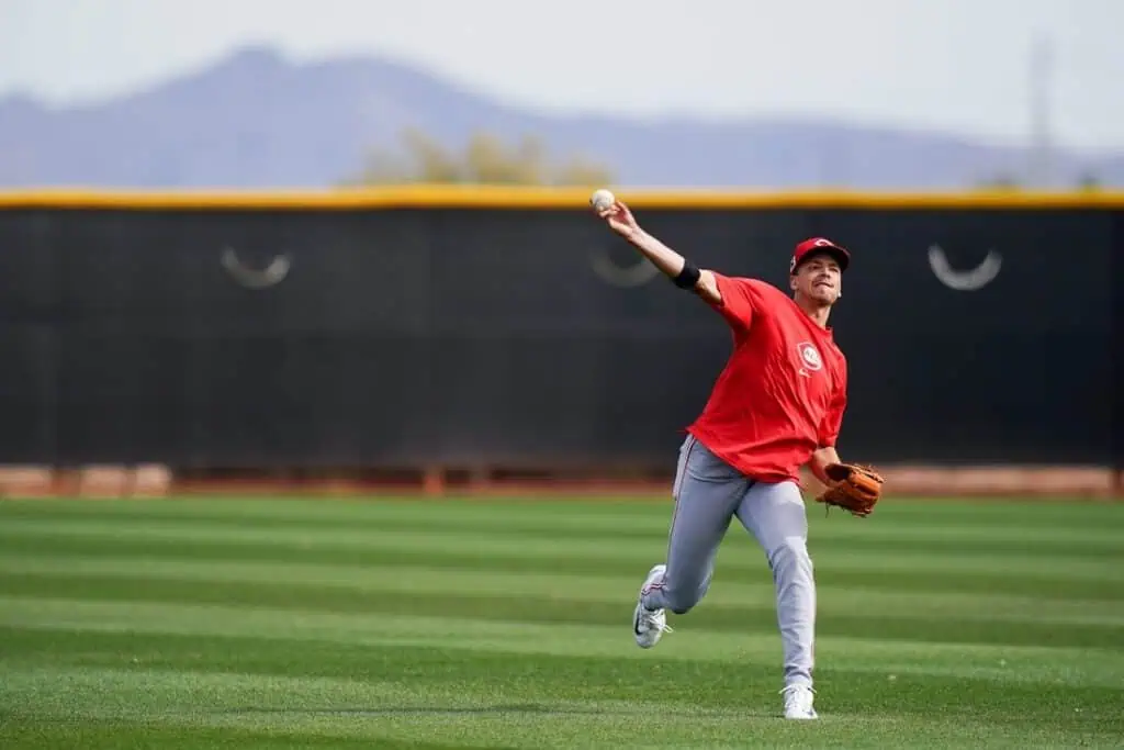 A baseball player in a red jersey throws a ball on a green field with a black backdrop