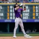 A baseball player in a purple jersey celebrates at first base, with a scoreboard displaying game scores in the background.