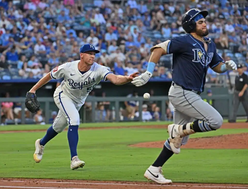 A Kansas City Royals player reaches for a baseball while a Tampa Bay Rays runner dashes toward first base in a lively baseball game.