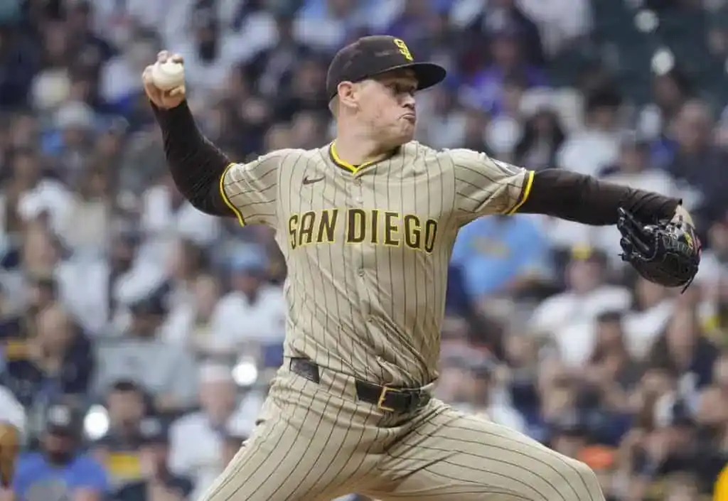 A San Diego Padres pitcher winds up to throw a baseball, wearing a tan and brown striped uniform, with a crowded stadium in background.