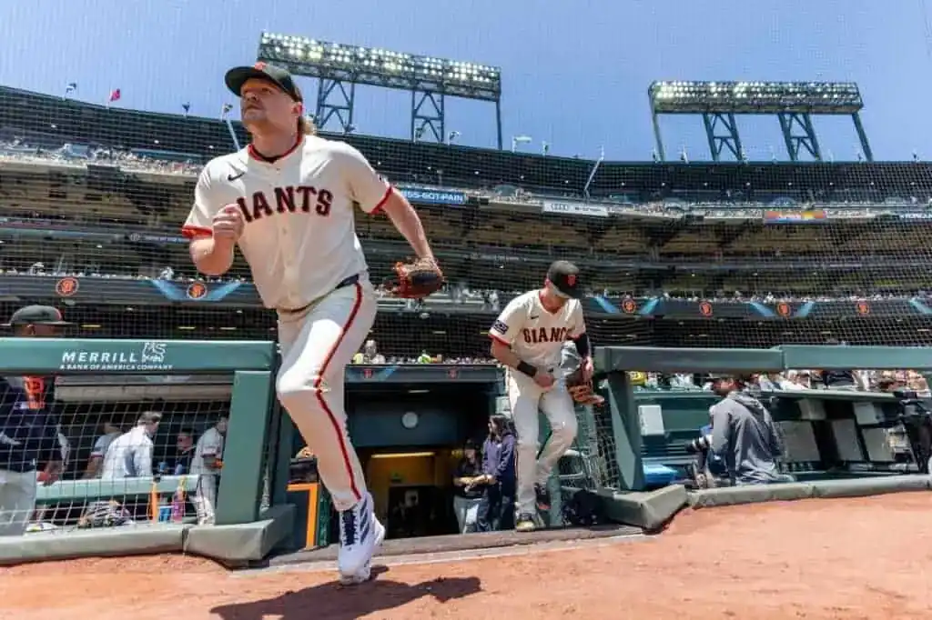 Two players in white San Francisco Giants uniforms run out from the dugout onto the field on a sunny day at the ballpark.