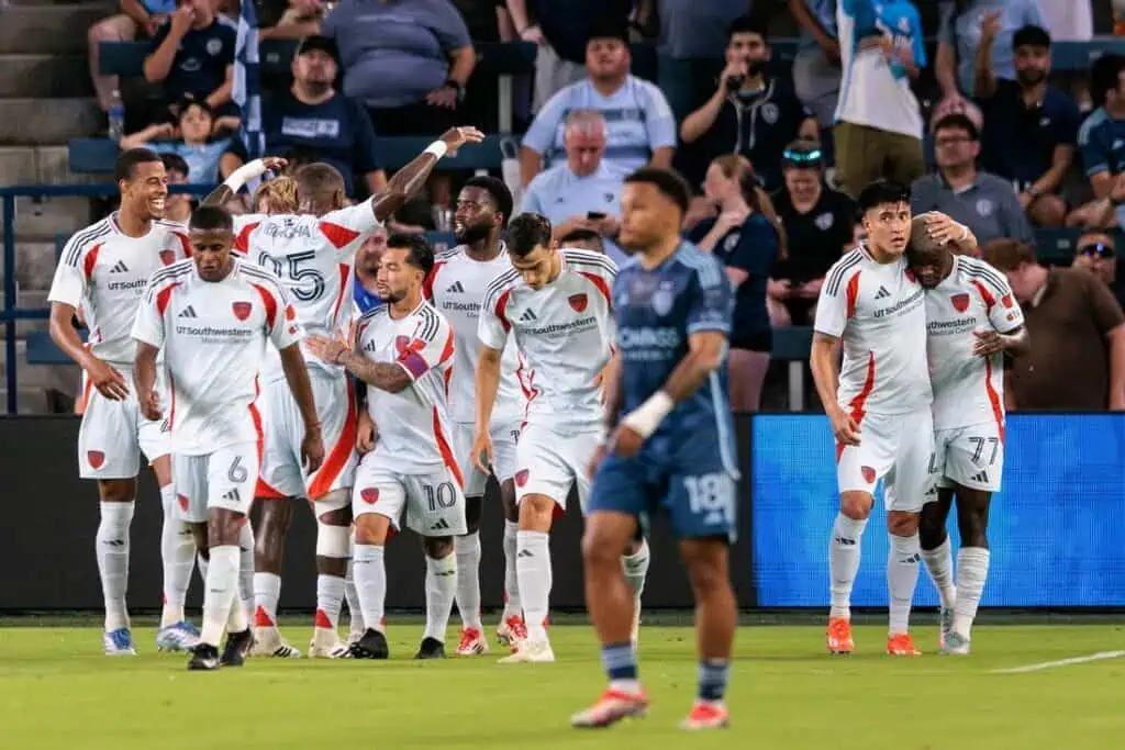 A group of soccer players in white jerseys celebrate a goal on the field, with fans cheering in the background.