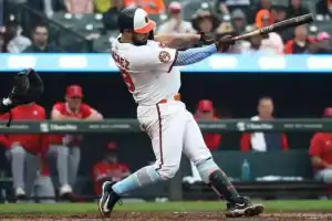 A baseball player in a white uniform swings a bat, preparing to hit a pitch, while teammates in red jerseys observe from the dugout.