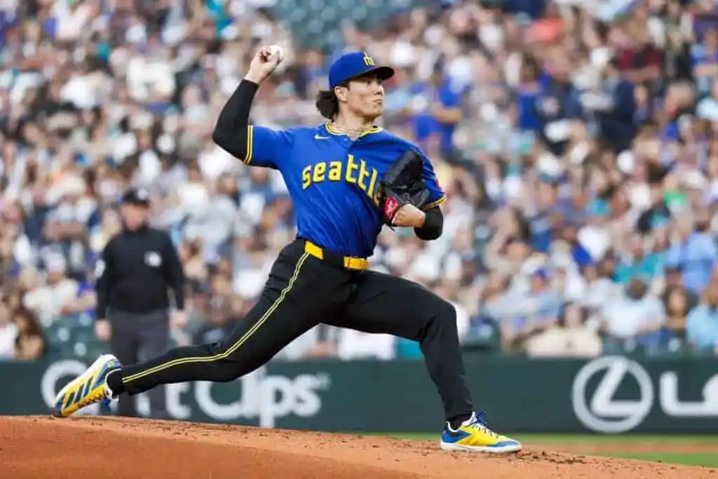 A baseball pitcher in a blue Seattle Mariners jersey winds up on the mound during a game, with fans in the background.