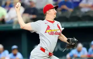 A pitcher in a gray St. Louis Cardinals uniform is winding up to throw a baseball during a game, with spectators in the background.