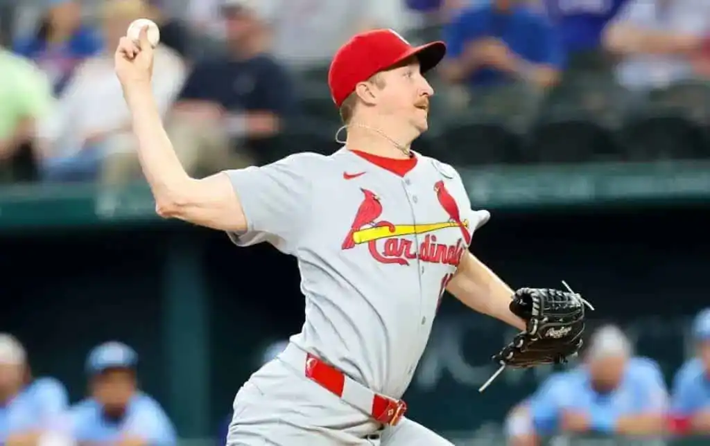 A pitcher in a gray St. Louis Cardinals uniform is winding up to throw a baseball during a game, with spectators in the background.