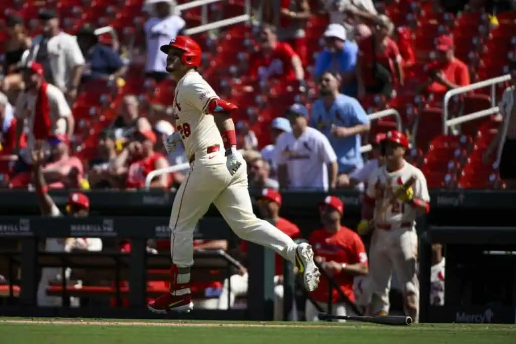A St. Louis Cardinals player runs past the dugout, with a crowd of fans in red jerseys cheering in the background at the stadium.