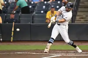 A baseball player in a white uniform swings a bat, aiming to hit a pitched ball during a game, surrounded by spectators.