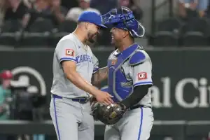 Two baseball players in gray uniforms converse on the field, one dressed as a catcher with blue gear. Fans are visible in the background.