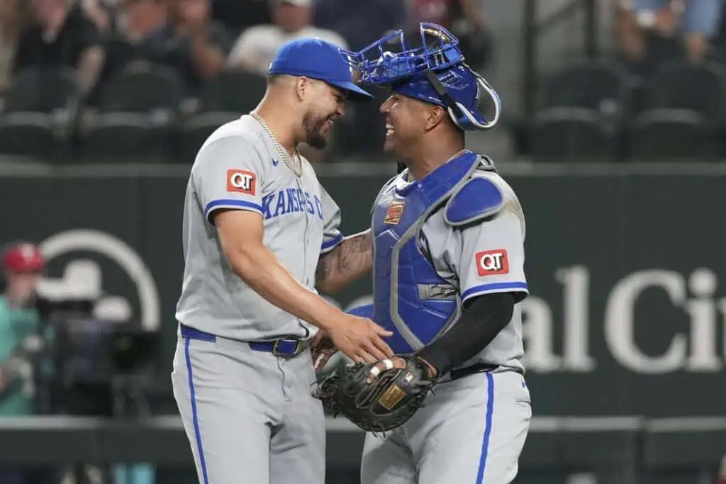 Two baseball players in gray uniforms converse on the field, one dressed as a catcher with blue gear. Fans are visible in the background.