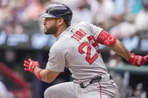 A baseball player in a gray uniform swings a bat, showing intense focus during a game. Spectators are blurred in the background.