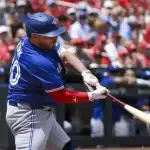 A baseball player in a blue jersey swings a bat during a game, surrounded by a cheering crowd in red and blue attire.