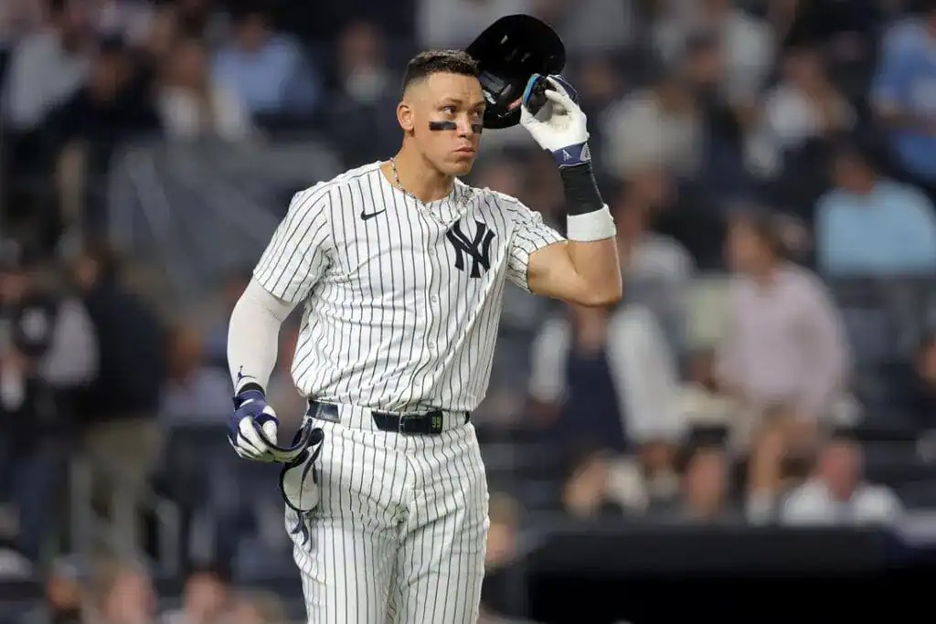 A New York Yankees player in a pinstripe uniform adjusts his helmet during a game, with fans blurred in the background.