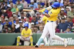 A Boston Red Sox baseball player, wearing a yellow City Connect jersey and blue helmet, is in mid-swing, making contact with the baseball. Spectators are visible in the blurred background.