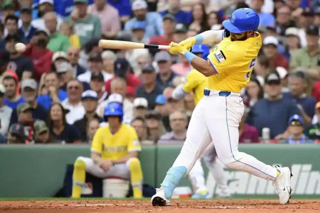 A Boston Red Sox baseball player, wearing a yellow City Connect jersey and blue helmet, is in mid-swing, making contact with the baseball. Spectators are visible in the blurred background.