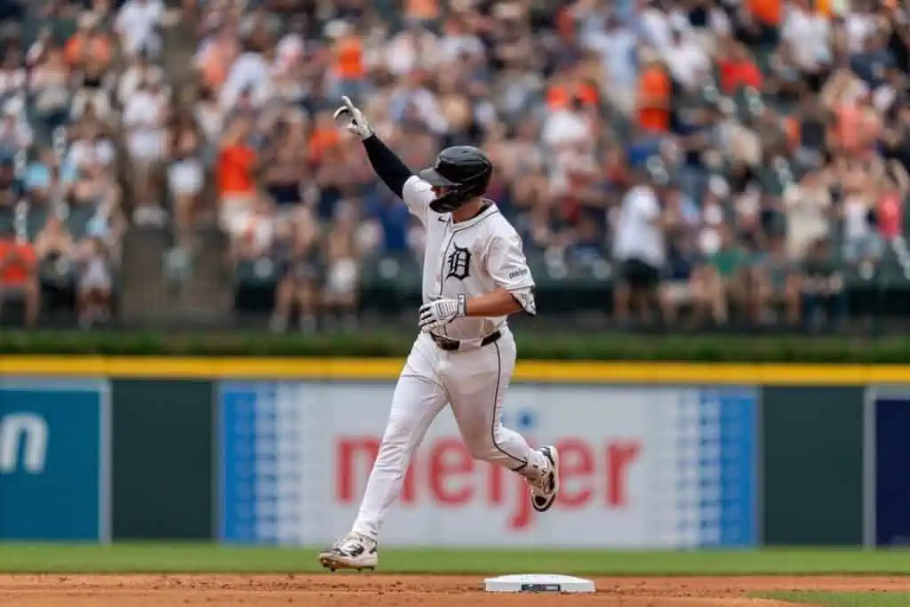 Detroit Tigers baseball player in a white uniform and black helmet running the bases and pointing upward during a game, with a blurred crowd in the background.