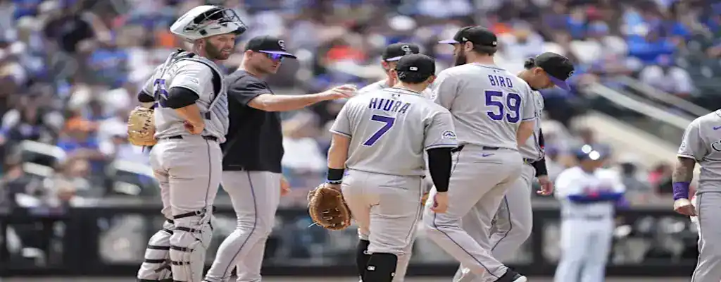 Colorado Rockies players gather on the mound during a game, wearing gray uniforms as they discuss strategy under sunny skies.