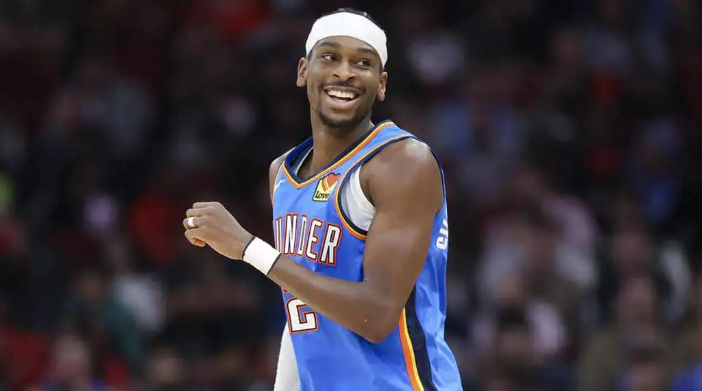 A basketball player in a blue Oklahoma City Thunder jersey celebrates on the court during a game, with fans blurred in the background.